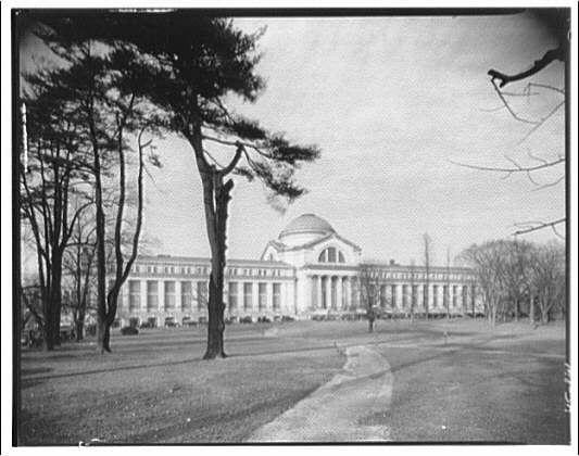 This black and white photograph depicts the historic Museum of Natural History, which is a large neoclassical building with columns supporting its portico. The structure has multiple floors and includes a prominent dome at one end that suggests it was built during an era when such architectural features were popular in institutional design. Trees surround the museum grounds, indicating that this photograph may have been taken on or near campus where such greenery is common for aesthetic appeal as well as environmental benefits like shade and air quality improvement. The absence of people gives a sense of quietude to what could typically be quite busy surroundings during exhibitions hours. There are no discernible signs or indicators giving away the exact location, time period beyond an early-to-mid 20th-century timeframe based on architectural styles referenced in provided information, nor any specific details within this image that would identify particular individuals if present at all times when it was taken.