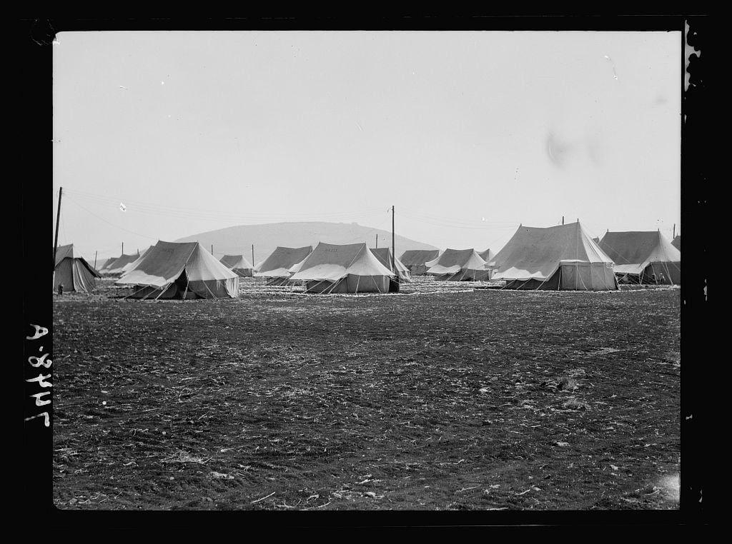 The image is a black and white photograph depicting several large tents spread out across an open field. The setting appears to be temporary, possibly for military or refugee purposes given the makeshift nature of the structures. In the background, there are hills which provide some depth to the scene.

In the lower-left corner of the photo, "A.S.P.U.T" is handwritten in capital letters, likely serving as an identification mark. The field itself appears barren and muddy with scattered debris or grass clippings on its surface. There's a lack of people visible within this photograph which adds to the impression that it was taken during a time when no individuals were actively using these facilities.

The photo is credited through metadata indicating "American Colony (Jerusalem). Photo Department", suggesting an affiliation with historical records from Jerusalem, likely documenting events or conditions related to American activities in the region. The specific mention of I.P.C., Iraq Petroleum Company camp and Tabor between 1934-1939 provides a contextual timeframe for when this photograph was possibly taken.

The overall composition is simple yet evocative; it captures an unassuming scene, seemingly frozen in time with no particular focal point. The monochrome palette enhances the historic atmosphere of the image without distraction from its content.