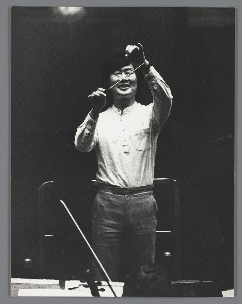 A black and white photograph depicting a person in casual attire, including trousers, sneakers, and a short-sleeved shirt. The individual is standing with one hand raised to the head level while holding onto what appears to be a conductor's baton or pole vertically above their left shoulder. They are smiling broadly towards the camera. There seems to be no musical context immediately apparent in this composition; it might capture an informal moment during preparation for performance, given the candid nature of the pose and expression.