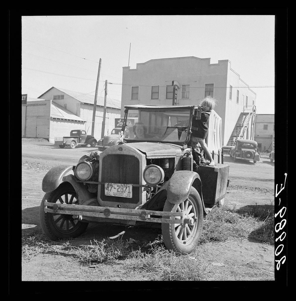 The image depicts a vintage automobile parked on an unpaved surface, with visible tire tracks and dry grass around it. The vehicle has distinctive features of early 20th-century design, such as round headlights, spoked wheels, and a large radiator grille. A person is standing next to the car, interacting with or possibly getting into it. In the background, there are other vintage cars parked on a street lined with buildings that appear industrial in nature. The scene suggests an older time period, likely several decades ago based on vehicle design and clothing styles of visible individuals.

The photograph's monochromatic color scheme indicates that this image is historical or styled to look as if it were taken during the early days of automotive travel when such vehicles would have been commonplace for daily use.