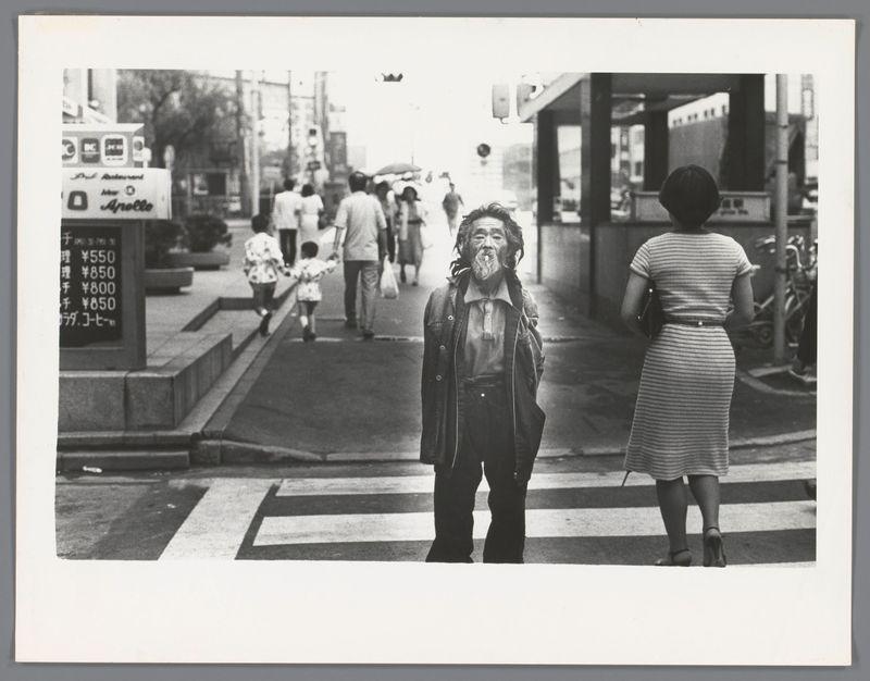 The image is a black and white photograph depicting an urban street scene. In the foreground, there's a man standing in the middle of what appears to be a pedestrian crossing with his back partially turned towards the camera. He has shoulder-length dreadlocks, wears a dark coat over a light-colored shirt, pants visible underneath the coat, and is holding something that looks like paper or documents near his waist.

To the right side of this man stands a woman facing away from the camera; she's wearing a striped knee-length dress with high heels. She seems to be in motion, perhaps walking.

The background shows pedestrians moving about on what appears to be a busy street. There are various storefronts and signs, including one that reads "Apollo" which might indicate an Asian business or district. A bus is partially visible at the far left edge of the image with Japanese text above it suggesting this scene could take place in Japan.

The overall mood seems candid and captures everyday life on a city street.