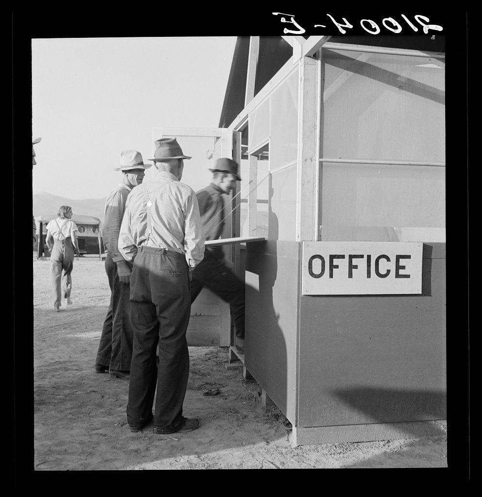 The black and white photograph captures a scene with four individuals gathered at an outdoor office setup. The structure is simple, with metal siding and a flat roof, marked prominently by a sign that reads "OFFICE." Two men are standing in line, one of whom appears to be interacting with someone inside the booth through a service window, while another man stands beside him, waiting his turn or possibly observing the interaction. A fourth individual is walking away from the office structure.

The setting seems arid and dusty, suggesting a rural environment that may have been typical for agricultural work in Oregon during the period when such structures were commonly set up to manage farm labor registration under programs like FSA (Farm Security Administration). The attire of the individuals—long-sleeved shirts, hats, and trousers—indicates casual or practical wear suitable for outdoor work. One man is wearing overalls.

The overall atmosphere conveys a sense of routine administrative activity possibly related to agricultural employment during that era.