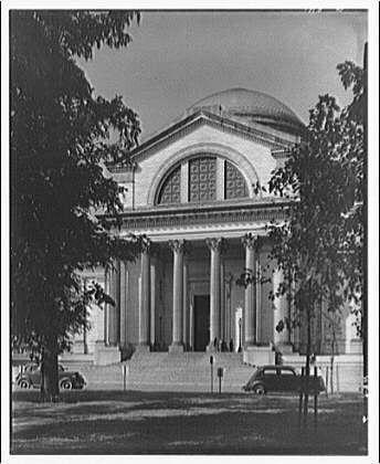 The image is a black and white photograph of the entrance to what appears to be an important public building, likely a museum or government institution. The structure has classical architectural elements such as columns supporting a pediment above the main entryway. There are trees framing either side of the building, and parked cars can be seen in front of it, suggesting that this photo was taken during a time when automobiles were commonly used but not yet ubiquitous.

The photograph is labeled "Entrance of Museum of Natural History ca. 1920-ca. 1950," indicating that the museum depicted here is likely to have been photographed around those dates. The label also provides additional information about the photographer, Theodore Horydczak (approximately 1890-1971), and notes a negative size of approximately "8 x 10 in."

The image appears on an online platform called Loener.nl, which seems to focus on historical photographs or art. The photograph's source is attributed as the Smithsonian Institution exteriors collection. This suggests that Horydczak may have been associated with this institution at some point during his career.

Overall, the photo captures a moment in time when urban architecture and transportation were evolving rapidly, likely illustrating how people interacted with public spaces and institutions like museums.