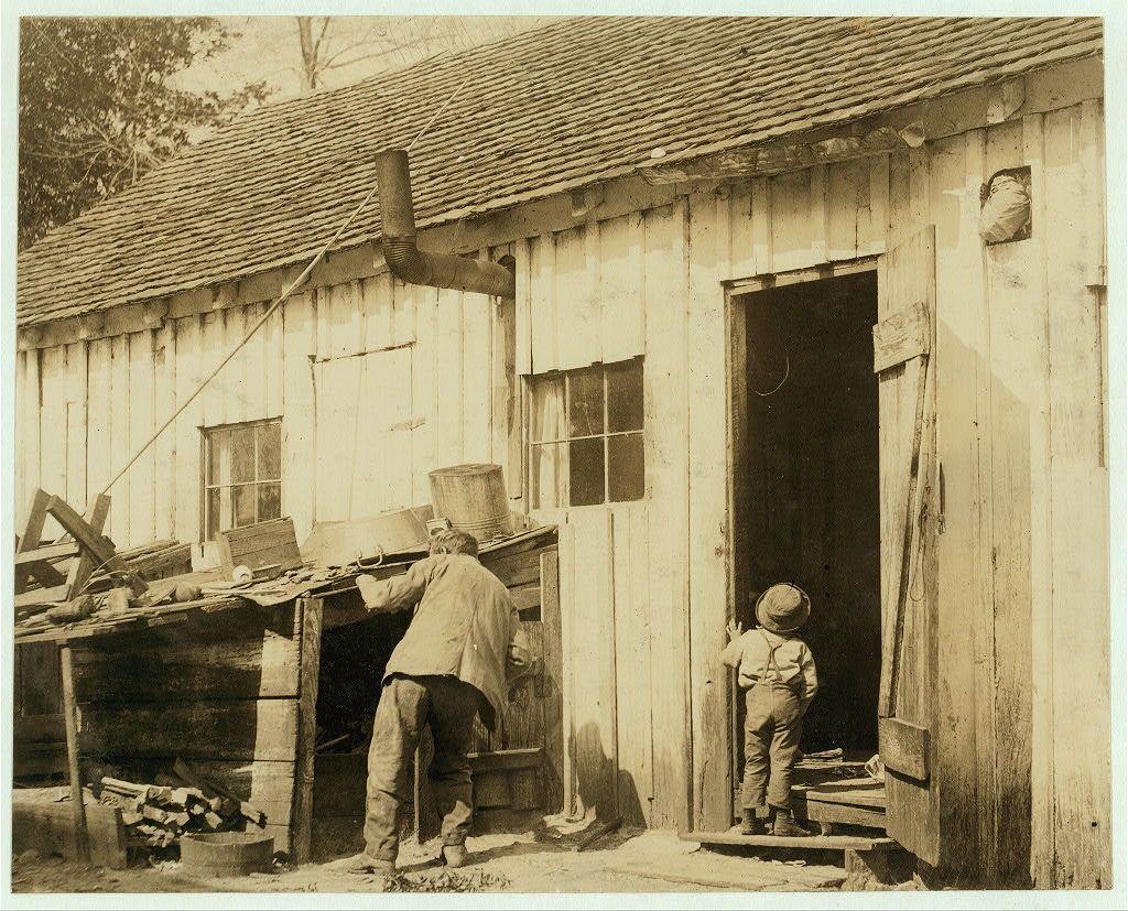 The image is a black and white photograph depicting an outdoor scene with two individuals near a rustic wooden building. One individual, seemingly an adult male dressed in work attire including trousers and possibly boots, stands inside the open doorway of what appears to be a shed or workshop. He's bending slightly towards something on his left side where piles of items like buckets are visible.

Adjacent to him is a child standing outside the door, observing the scene with interest. The child wears overalls with suspenders, suggesting practical clothing for outdoor work or play. There's an assortment of objects around them including stacks of wood and possibly tools scattered near their feet on what looks like soil ground, hinting at a rural setting.

The structure shows signs of wear and age with its weathered walls and slanted roof covered in shingles. A chimney is visible to the left side indicating some form of indoor heating or cooking activity might take place within this dwelling. The surrounding environment seems sparse aside from the building itself which implies a modest living condition, possibly reflective of working-class housing.

The sepia tone indicates that it's likely an older photograph, perhaps dating back over a century considering its quality and style. There is no visible text indicating any specific narrative or context beyond what has been described.