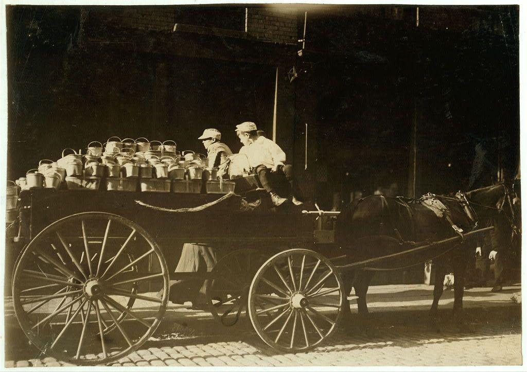 The image depicts a historical scene featuring an old-fashioned horse-drawn wagon loaded with metal cans, possibly used for transporting goods. Two individuals dressed in period clothing sit atop the pile of containers on what appears to be a cobblestone street or market area. The person closer to the camera is wearing a white hat and jacket, while the individual further back wears a darker outfit. A horse stands behind the wagon, equipped with harnesses but not actively pulling it at this moment.

The photograph has an aged appearance, consistent with early 20th-century imagery, suggesting that it may capture daily life or work in urban settings of that era. The black and white composition emphasizes contrasts between light and shadow, adding to its vintage feel.
