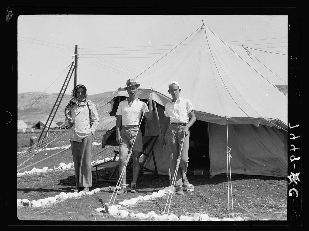 The image depicts three individuals standing outside a large white tent in what appears to be an arid, outdoor setting. The person on the left is wearing traditional Middle Eastern attire, including a headscarf and long robe with trousers, holding ski poles or sticks. In the center stands another individual dressed in light-colored western-style clothing, complete with shorts, sandals, and a hat, also armed with similar sticks held upright to their body height. To his right, there's an additional figure wearing white clothes including gloves and shoes that resemble those of European origin from around the 1930s. The tent has a conical shape typical for desert or military-style camps during exploration expeditions in such regions.

The background reveals more tents secured on poles with ropes, hinting at a temporary settlement, perhaps associated with an exploratory campsite as suggested by the context provided through the caption mentioning "Iraq Petroleum Company" and "American Colony (Jerusalem)" along with "Photo Department". The environment is characterized by rocky terrain without vegetation, indicative of desert conditions. There's a clear sky above them which could suggest it’s either morning or late afternoon given the quality of light.

This photograph seems to capture workers from different cultural backgrounds possibly involved in petroleum exploration work aro [...]