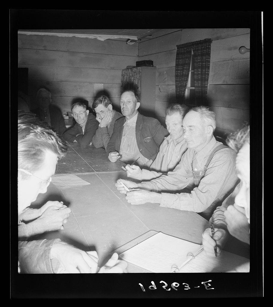 The image is a black and white photograph depicting several men seated around a table in what appears to be an indoor setting with wooden walls. The individuals are engaged in discussion, some leaning on the table while others hold papers or pens, indicating they might be involved in deliberation or decision-making processes.

On the left side of the frame, part of another person's head is visible but not fully included within the image. This suggests that more people may be present outside the captured area. The men are dressed casually; some wear jackets and others appear to have on work attire suitable for manual labor. Their expressions range from contemplative to engaged.

The setting seems informal, possibly a community meeting or council discussion among rural workers. A window with curtains is visible in the background, providing natural light that illuminates part of the room. There's no direct indication of technology like computers; instead, it suggests an older time period based on attire and interior design elements.

The image has some textual information at the bottom: "13205-E," which could be a cataloging or identification number related to its source collection (e.g., Farm Security Administration photo). The context provided by this reference hints that the photograph is part of an archive concerning farm labor conditions, possibly during mid-20th-centur [...]