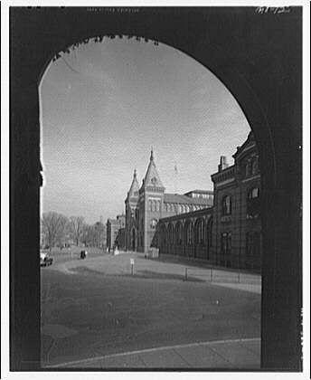 The image shows a vintage black and white photograph of an archway leading to the Smithsonian Institution Building, also known as The Castle. This iconic structure is prominently visible through the opening of the arched passageway in what appears to be Washington D.C., based on its architectural style.

In the background, there are several buildings with distinctive features such as pointed roofs and large windows that suggest a historical significance or institutional use. A few cars can be seen parked outside The Castle's grounds, indicating it was taken during an era when automobiles were already in common usage but not yet ubiquitous.

The sky is clear with no visible clouds, suggesting fair weather conditions at the time of the photograph. There are bare trees around which hint that this image could have been captured either late fall or winter season as there’s a lack of leaves on them.

Overall, the photo provides an atmospheric view into early to mid-20th-century Washington D.C., with The Castle serving as its central focal point and historical landmark.
