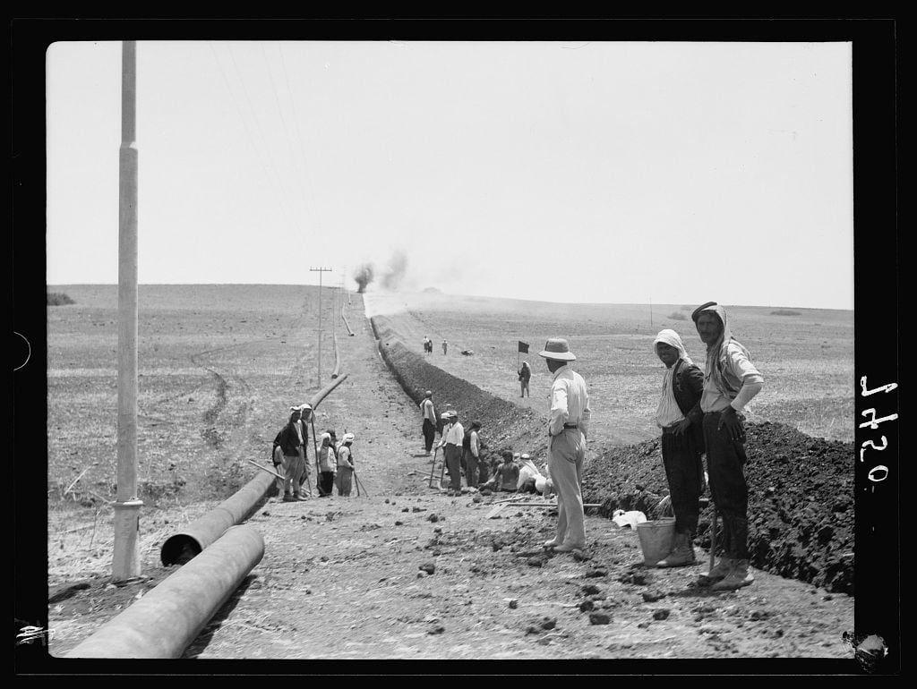 The image depicts a historical black and white photograph of workers engaged in pipeline construction. The setting appears to be an arid, open landscape with sparse vegetation under clear skies. A long horizontal pipe is laid on the ground along what looks like a trench or prepared earthwork.

In the foreground, several individuals are seen working around this infrastructure project; some standing while others sit or lie down. Some of them wear wide-brimmed hats and headscarves, suggesting protection from sunlight. One person stands with hands in pockets near a bucket on the ground, possibly containing tools or materials for work.

A group is gathered along one side where construction seems to be concentrated, showing activity around what appears to be an explosive blast site indicated by smoke rising into the air. The workers' casual postures and relaxed demeanors suggest they might not have been directly involved in this particular action but are observing it from a distance.

The scene captures a moment of laborious work during the era suggested by historical context, possibly related to the oil industry or infrastructure development projects such as those conducted by companies like Iraq Petroleum Company (IPC). The photograph is labeled with "J120" and credited to American Colony (Jerusalem), which indicates its source.