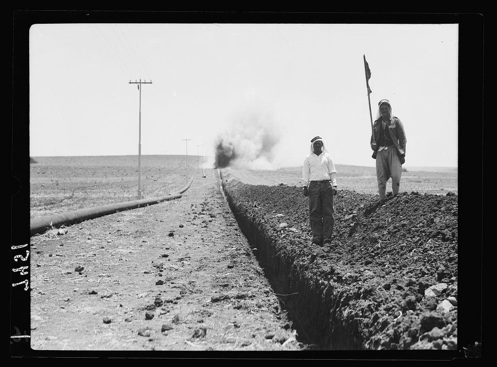 The image is a black and white photograph depicting two individuals walking along the edge of an excavation trench in what appears to be arid, open terrain. One person holds a flag or banner aloft while carrying tools that suggest they may be working on constructing or repairing something underground, possibly related to pipeline work given the visible large pipe laid alongside them.

In the background, there is smoke rising from somewhere within this vast expanse of land, hinting at some activity taking place farther away. The environment appears barren with a lack of vegetation and only sparse structures like telephone poles punctuating the landscape in the distance.

The photograph has been cataloged as being taken by I.P.C., possibly relating to the Iraq Petroleum Company during their operations between 1934 and 1939, according to information provided. The photo is labeled with a negative label or title on its border that reads "Preparing trench for pipe line on Esdraelon," which suggests this scene might be part of an archaeological dig related to the Esdraelon region.

The image has been attributed to American Colony in Jerusalem's Photo Department, indicating it may have historical significance and was possibly captured as a record of activities during a specific time period.