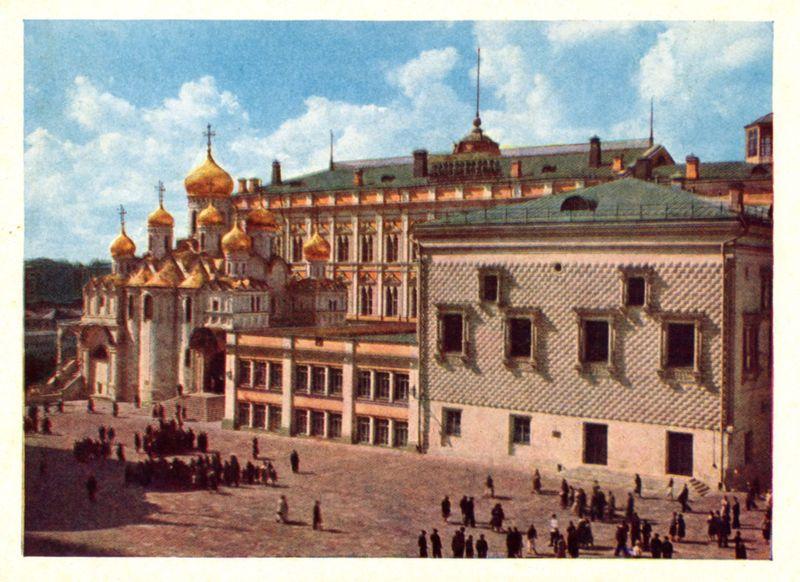 The image depicts a historical scene of the Kremlin in Moscow, Russia. The focal point is an ornate building with multiple golden domes and crosses atop its towers, indicative of Russian Orthodox architecture. This structure stands prominently against a backdrop of blue sky dotted with white clouds.

In front of this edifice lies a spacious square bustling with people engaged in various activities—some walking individually or in small groups, while others appear to be gathered around particular points of interest on the pavement. The ground is paved, and there are several individuals sitting on what seems like steps leading up to the building's entrance.

To the right stands another imposing structure with a simpler architectural design featuring regular rectangular windows, some adorned with curtains or blinds partially drawn down. This side contrasts with the ornate façade of the Kremlin but adds balance to the overall composition.

The image conveys a sense of everyday life in Moscow during an era that appears somewhat historical based on clothing styles and architecture. The colors are vivid, with warm hues dominating due to sunlight bathing the buildings, while hints of cooler tones punctuate from patches of sky visible around the edges of the frame.