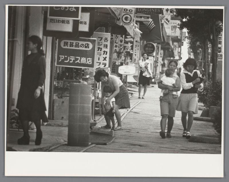 The image is a black and white photograph depicting an urban street scene. There are several individuals walking or engaged in activities along the sidewalk, which features various shops with signage written in Japanese characters. The people vary from adults to children; some carry items such as handbags, backpacks, and infants.

One woman appears focused on a task involving a cylindrical object at her feet, possibly cleaning or inspecting it. A child stands beside this adult figure, observing the activity closely. In another part of the image, two women walk together carrying an infant between them; they seem to be in conversation while walking down the street.

The shop signs include names and what appears to be product offerings like "Cookies" (possibly a translation error as it is not written clearly), indicating the presence of food-related businesses. The overall atmosphere suggests everyday life with people going about their daily routines on this city sidewalk, which may offer insights into social interactions or local culture during the era when the photo was taken.

The photograph's composition captures candid moments and provides glimpses into a slice-of-life scenario from Japan at that time period, as evidenced by Ed van der Elsken’s work.