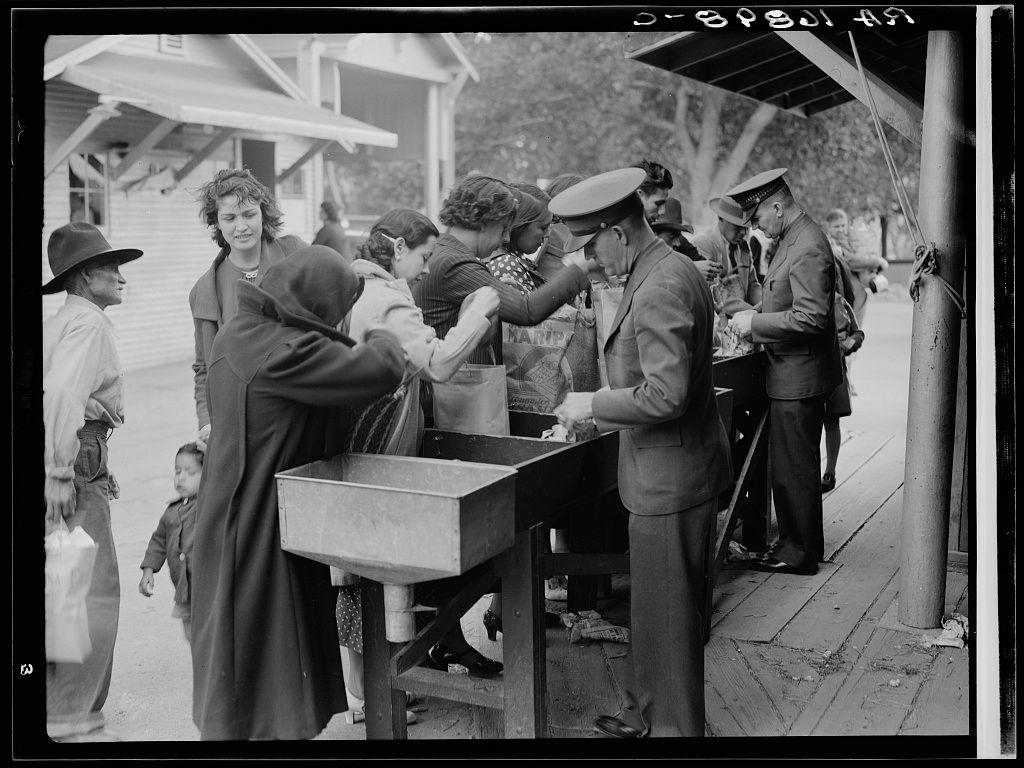 The image depicts a black and white scene from what appears to be the early 20th century, as indicated by the clothing styles. It captures several individuals engaging in various activities on a street or pavement setting with buildings visible in the background.

There is an older man wearing military-style uniform handling items at a makeshift stand where he seems to be handing out food and drinks from containers onto paper bags held by women who are receiving them, suggesting some form of relief distribution. Another woman stands near him, possibly waiting for her turn or overseeing the process, with another child close by.

A group of people is gathered around in casual conversation or interaction with each other; a mix of adults and children can be seen among them. One man on the left wears a wide-brimmed hat while carrying what appears to be produce in bags over his shoulder.

The overall setting suggests a scene from an era of economic hardship, possibly during or after World War I when relief efforts were common due to unemployment rates leading up to and following the war period known as the Great Depression. The image carries historical significance related to American life during challenging times such as post-war recovery periods where government aid was distributed among affected populations.

The photograph is credited with being taken on June 29, 1940, by D [...]