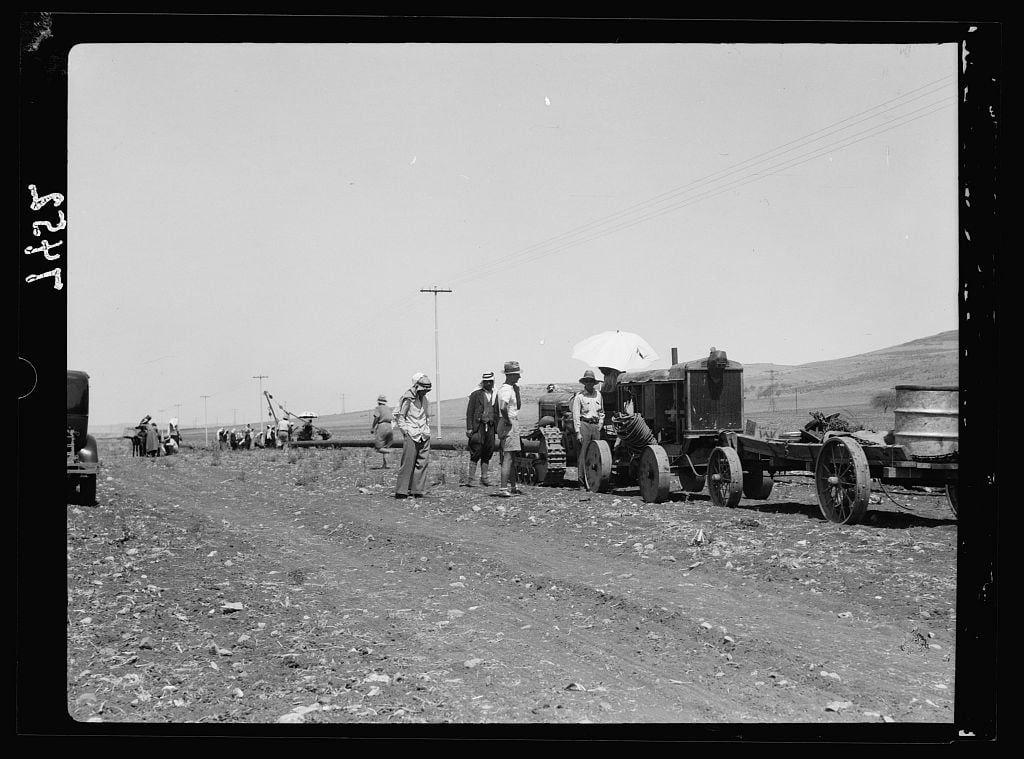 The black and white photograph captures a group of men working outdoors, likely in the 1930s. The scene depicts rural or undeveloped terrain with an unpaved dirt road stretching into the distance under clear skies. Several individuals are gathered around old-fashioned farming equipment: tractors, a plow attached to one tractor, and various carts carrying what appears to be agricultural tools.

The men wear hats for sun protection, and some carry umbrellas possibly indicating bright or harsh sunlight. The group is organized in small clusters, with one man seated on the ground, suggesting rest or waiting time during work intervals. In the background, additional farming equipment can be seen farther down the road along with a utility pole.

The overall atmosphere of the image suggests agricultural labor, perhaps related to irrigation projects or general land preparation for crops. The absence of modern machinery indicates that this was taken in an earlier era when such manual and rudimentary methods were common.