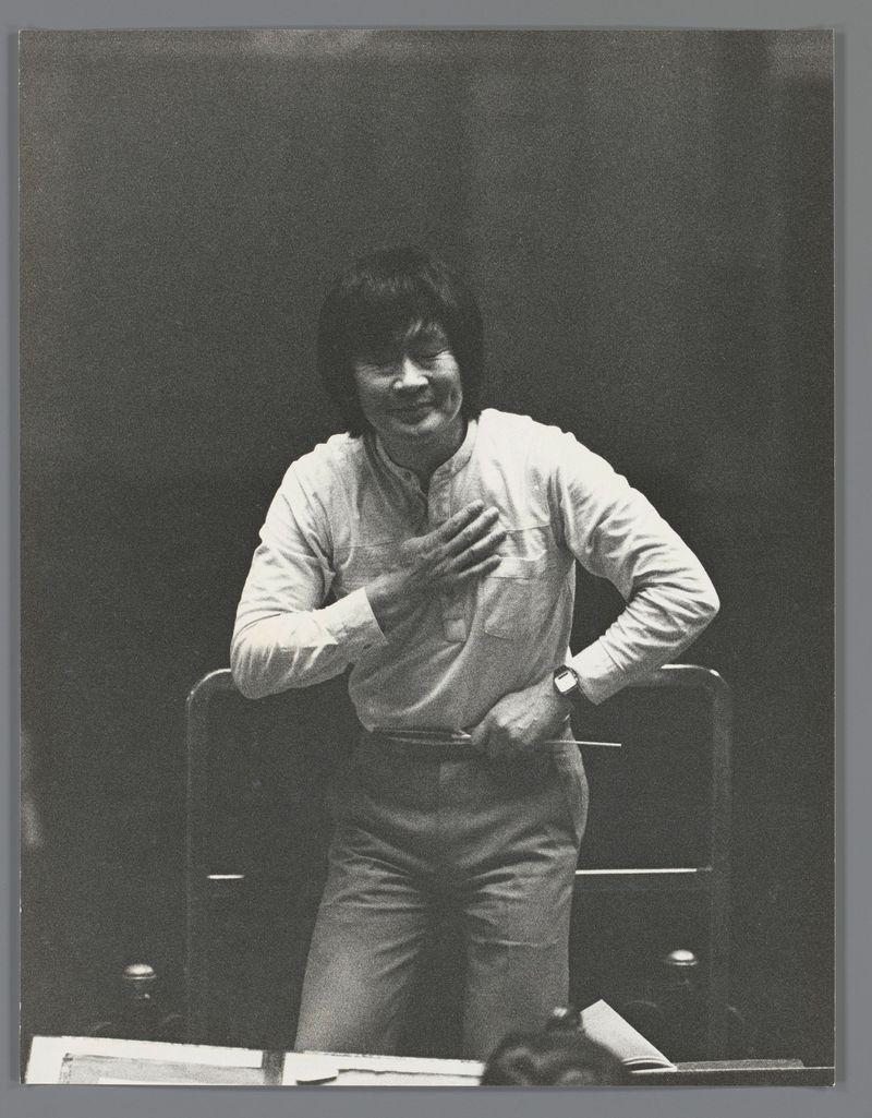 The image is a black and white photograph of an individual who appears to be in the middle of conducting. The person has dark hair, wears a light-colored long-sleeved shirt with rolled-up sleeves, khaki trousers, and dress shoes. They are standing behind what seems like a podium or music stand that holds books or sheet music. Their left hand is placed on their chest while their right arm extends forward as if gesturing to an orchestra. The individual has a watch visible on the wrist of this extended arm.

The background is dark, focusing attention on the subject and creating a contrast with their lighter clothing. No clear facial features are discernible due to blurring or distance from the camera lens. There's no additional context provided about the location or event taking place in the photograph itself.
