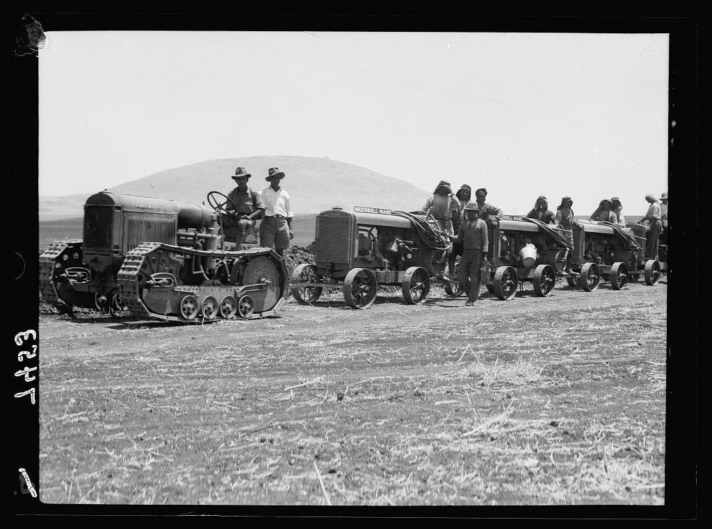 The image is a black and white photograph showing several tractors lined up in an outdoor setting. The terrain appears to be dry with sparse grass, indicative of arid conditions or drought-stricken land. In the forefront stands one large tractor equipped with tracks instead of wheels for better traction on uneven ground. On this vehicle are two individuals wearing hats typical of early 20th-century western attire; their presence suggests they may be operators or crew members.

Behind it is a longer line-up consisting mainly of older-fashioned, non-track tractors that have large front grilles and prominent radiator caps, indicative of the era's design. The people associated with these vehicles are seen in various poses: some standing atop, others beside them holding hoses connected to what seem like fuel containers or tanks.

The backdrop features a hazy skyline with mountains visible on the horizon under an overcast sky. There is no discernible movement captured within this still photograph; it captures a moment of calm and organization as if these vehicles are part of a convoy for transportation, work purposes in agriculture, or another collective endeavor from that period's socioeconomic context.

The presence of individuals dressed similarly to ranchers suggests they might be farmers or workers belonging to an agricultural company. There is no explicit indication what  [...]