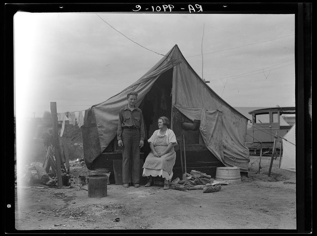 The image is a black and white photograph depicting two individuals, presumably a man and woman, standing in front of a makeshift tent or shack. The structure appears to be constructed from various materials including canvas for the walls and some tarpaulin fabric on top. There are personal belongings scattered around them such as buckets, tools, and clothes hung out to dry. In the background, more similar tents can be seen, suggesting an encampment of individuals possibly in a transient or difficult living situation.

The man is standing with his hands behind his back wearing what appears to be work clothes including boots. The woman sits on a low stool beside him dressed in simple attire that includes a skirt and shirt, sitting cross-legged as she looks towards the camera. Both individuals have solemn expressions on their faces which may convey hardship or fatigue.

The setting seems to be an outdoor area with dirt ground, indicative of rural or unimproved urban conditions during what appears to be daytime based on natural lighting in the photo. There's a handwritten text visible at the top left corner that reads "9-10PP - A8," which could indicate a cataloging system used by photographers like Dorothea Lange for her work.

The image captures a moment of their daily life during what seems to be tough economic or social conditions, possibly related to rural labor migrati [...]