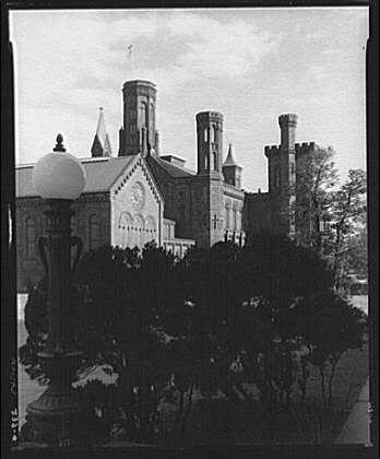 The image depicts a historical building with architectural features reminiscent of Romanesque Revival or Gothic styles. The structure has multiple towers and spires, suggesting it may be a church, cathedral, or another significant religious edifice. In the foreground stands a street lamp with a classic design featuring an ornate base and a large, round light bulb on top. Trees are visible around the area, partially obscuring the building's facade but providing some context for its setting as likely being in a park-like environment within a city. The photo is presented in black and white, which gives it a timeless quality and emphasizes contrasts of light and shadow across the scene.