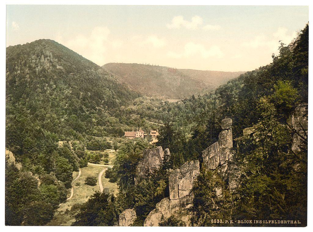 This image depicts a picturesque landscape view from the Hartz mountain range in Ilfeld, located within Germany. It showcases an expansive valley with lush greenery covering rolling hills and dense forests. A prominent feature is a rocky cliffside adorned with vegetation and scattered boulders that provide a stark contrast to the surrounding verdant terrain.

A narrow winding road meanders through the landscape, leading towards what appears to be a small rural settlement or estate nestled among trees. The architecture of this building seems traditional, possibly indicative of historical significance in the area. Above it all is a partly cloudy sky with soft hues that complement the natural beauty below.

The image has an aged look, suggesting it may have been taken during the late 19th to early 20th century. There's text at the bottom right corner which reads "8536 P.E.: BLICK INS ILEFSDERHAL", likely a reference or cataloging information pertaining to this photograph.

The overall scene exudes tranquility and highlights the scenic charm of rural Germany during that period, inviting viewers into an idyllic landscape where nature's splendor is on full display.