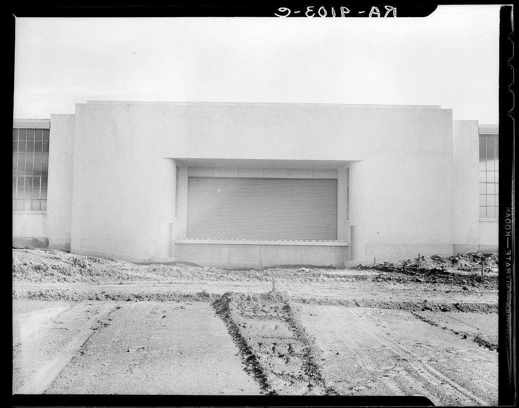 This black and white photograph depicts a stark, industrial setting. The main subject is the side facade of an unfinished or abandoned building with exposed concrete walls. A large, rectangular window shutter dominates the center of this structure's wall. It appears weathered, hinting at its age and exposure to harsh elements.

The ground in front of the building shows evidence of recent activity, as evidenced by tire tracks etched into a dirt surface that looks like it has been raked or smoothed over with some sort of implement. These marks lead up to what seems to be an entrance point for vehicles, which could suggest regular use despite its state.

To one side is another structure, perhaps part of the same complex; however, only parts are visible in this shot - a series of windows and a portion of a wall that also show signs of wear. The absence of human presence adds to the sense of abandonment or disuse.

The photograph's monochromatic palette enhances its starkness and could be indicative of it being taken during times when color photography was less common, such as early to mid-20th century America - which is possibly relevant given the reference to a Hightstown New Jersey factory for garment workers.