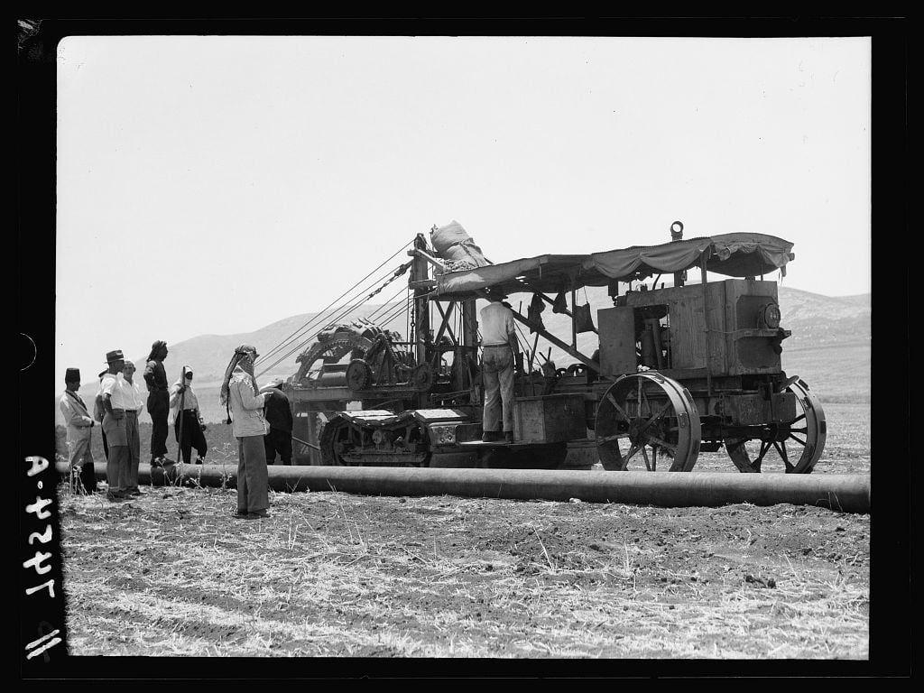 The image depicts a historical scene with multiple individuals and machinery in what appears to be an agricultural or construction context. In the foreground, there is a large cylindrical object lying horizontally on the ground which looks like a pipeline or irrigation pipe. Several people are standing around this object; some seem to be workers while others might be overseeing the operation.

In the middle of the scene stands an old-fashioned tractor with multiple attachments and components, including what seems like a drilling apparatus mounted on top. The tractor has large wheels and is connected via cables to various parts of the machine being operated or tested by one individual standing atop it. There are also some fabric coverings draped over certain sections of this setup.

The background shows an open field with sparse vegetation under a clear sky, suggesting that this activity might be taking place in rural areas or on undeveloped land. The people around have varying styles of clothing and hats indicative of different roles within the team; for instance, one individual wears what looks like military-style uniform while others wear more casual attire.

Overall, this image captures an early 20th-century scene likely associated with oil exploration or pipeline construction activities in a region under British control during that era.