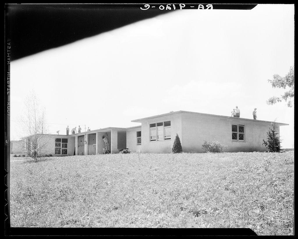 The image is a black and white photograph showing an expansive field with tall, dry grass. In the background, there's a single-story house characterized by its rectangular shape, flat roof, and large windows. The structure appears modern for its time, indicative of mid-century design aesthetics that emphasize simplicity and functionality. A series of steps lead up to one side entrance where two individuals are standing, suggesting an interaction or conversation taking place outside the dwelling.

The sky is clear with minimal cloud cover, indicating fair weather conditions. There's a tree visible on the right-hand side at a distance from the house, adding to the rural feel of the setting. The grassy field extends into the foreground, occupying most of the frame and creating a sense of openness around the building. No other structures or significant landmarks are in view.

The photograph has text overlaying it with white lettering against the black background at the top corner reading "9-061P-B2," which could be an archival reference number for this image. At the bottom left, there's another line of alphanumeric characters that read: "GODENWERT - FRA[...]", likely a part of a longer caption or title associated with this photograph.

The inclusion of people in front of the house gives life to the otherwise static scene and provides context regarding scale and use. The indiv [...]