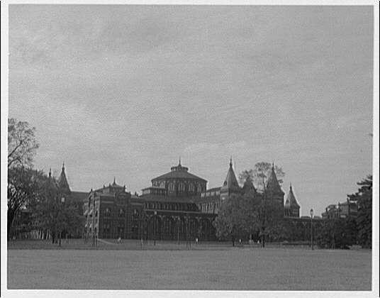 The image depicts an old, historic building with multiple towers and spires, suggestive of a Gothic architectural style. The photograph is in black and white, indicating it was likely taken during the early to mid-20th century based on its monochrome tone.

In front of this grand structure stands what appears to be a large open field or lawn, implying that the building might have been part of an expansive campus such as a university. The trees are sparse with autumnal colors; their leaves seem not fully developed yet suggest it may be early in fall season when most deciduous trees begin to lose their foliage.

There's no sign of modern technology like computers or cars which suggests this photograph was taken before the widespread use and prevalence of these items during that period. The sky is overcast, giving an impressionistic feel to the scene with a lack of shadows on buildings' facades suggesting either early morning or late afternoon lighting conditions.

The absence of people in such large spaces could suggest two things: it's not peak visiting time for this place (possibly due to its location being outside regular tourist hours), and there were fewer public gatherings at that era. Overall, the image evokes a sense of historical significance with an atmosphere indicative of a bygone era.