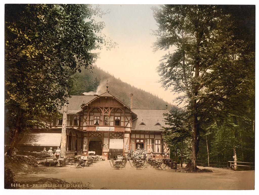 This image depicts a quaint, rustic inn with traditional timber-framed architecture in the German town of Ilsenburg. The building is adorned with "Prinzess Insel" signage on its facade and features several large windows that allow natural light to enter. A chimney from which smoke is billowing out suggests an active fireplace within. Surrounding the establishment are outdoor seating arrangements, including benches and tables where patrons can enjoy their meals or beverages amidst a serene forested setting. The lush greenery of trees frames the scene on both sides, indicating a tranquil environment away from urban hustle. In the background, a hill partially shrouded in mist adds to the picturesque atmosphere of this historical German locale dating between circa 1890 and around 1900.