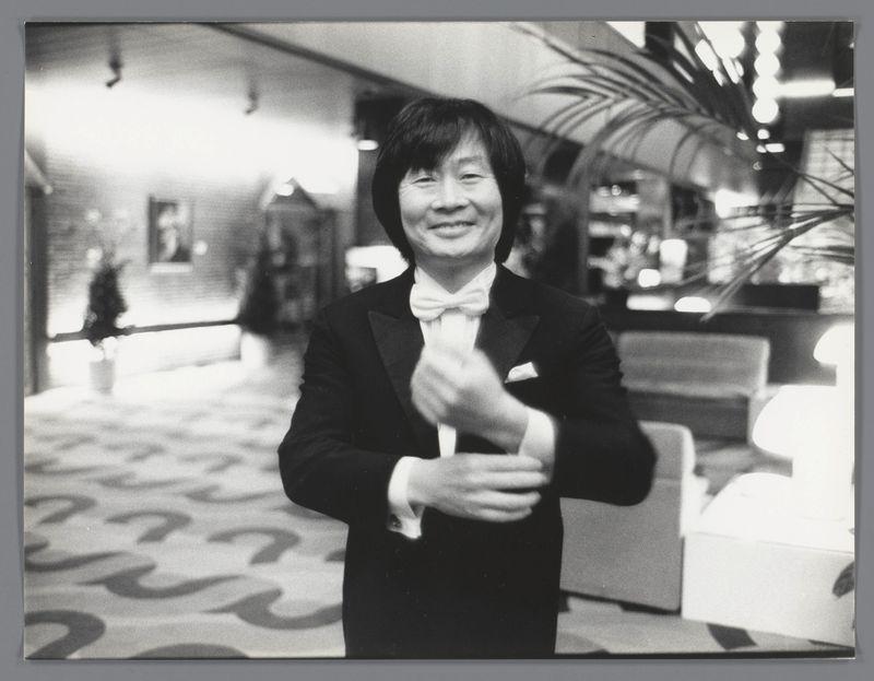 A black and white photograph depicting an individual wearing a formal suit with a bow tie. The person appears to be posing confidently, smiling directly at the camera while clutching what seems like a handkerchief or napkin in their right hand. They are situated indoors, likely within a public venue such as a theater lobby or hotel entrance hall, evidenced by seating arrangements and lighting fixtures visible in the background.