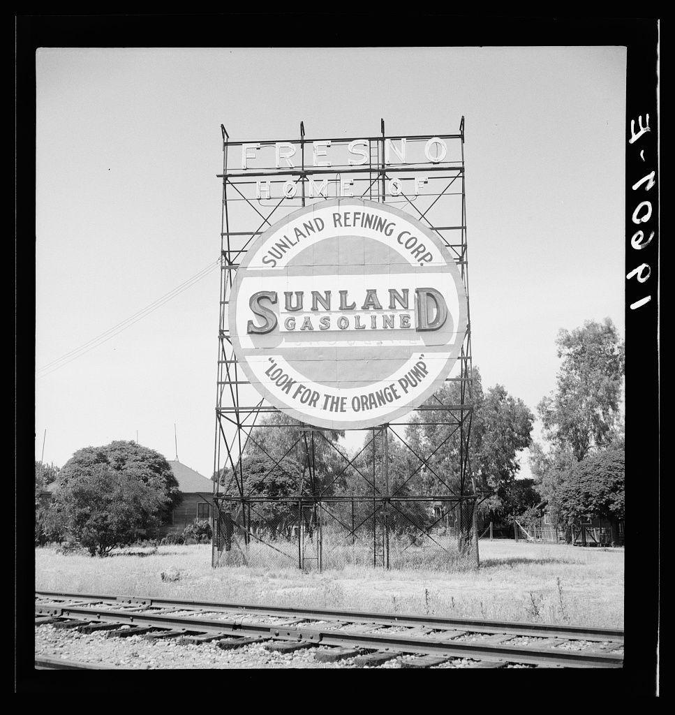 This black and white photograph captures a tall, metallic billboard for the Sunland Refining Corp., advertising their gasoline with an invitation to "Look For The Orange Pump." It stands prominently against a clear sky. In the foreground, there is evidence of railway tracks running parallel along the bottom edge of the frame, suggesting this sign might be located on or near railroad property in California's rural landscape.

The surrounding environment appears somewhat barren and desolate with sparse vegetation, including trees and shrubs scattered around. A modest structure can also be seen in the background, indicating human habitation nearby but not much activity is evident from these structures' appearance.

Given this context, it seems like a typical scene one might find on outskirt highways or rural areas of California during an earlier period when such gas station advertisements were common and prominent. The clear sky with no clouds hints at favorable weather conditions for travel and business activities associated with the advertised gasoline service.