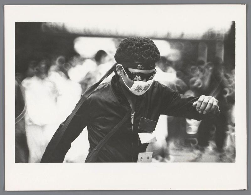 The image is a black and white photograph depicting an individual in motion, possibly walking. The person appears to be wearing traditional Japanese attire, including what looks like a martial arts uniform or a similar style garment, with prominent shoulder armor pieces known as 'dobok' visible on the shoulders.

Their face is partially covered by a cloth mask featuring some writing, which could indicate it's used for anonymity in public settings. The individual has curly hair and their right arm is extended forward, suggesting movement through a crowd or busy street environment.

The background is out of focus with several indistinct figures that resemble other people, likely pedestrians given the urban setting implied by this context. There are no discernible features indicating specific locations within Japan due to the lack of clear landmarks in the image provided.

Given the attire and mask, it's plausible this photograph could be connected to a cultural event or demonstration where such clothing is worn for symbolic reasons while maintaining privacy from identification.