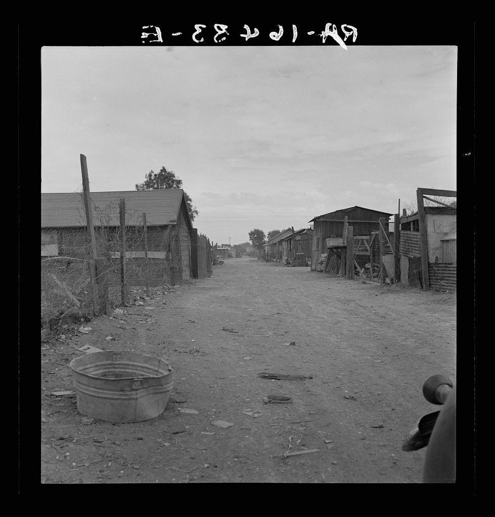 The image depicts a desolate urban environment, specifically showing makeshift and deteriorated housing conditions. A row of small wooden shacks with various degrees of disrepair lines one side of an unpaved road or alleyway. The structures appear rudimentary, lacking in modern amenities such as electricity or running water. In the foreground is a large metal basin on the ground, indicating potential use for collecting rainwater or other resources scarce within this community.

The sky above is overcast with no visible vegetation nearby, suggesting an arid climate typical of desert regions like Imperial Valley in California where field workers reside. The overall atmosphere conveys neglect and hardship associated with impoverished living conditions faced by Mexican immigrants working on the farms of the region during a time when they were subject to exploitation.

Historical context reveals that these slums represented common housing for agricultural laborers, highlighting social injustices such as inadequate sanitation facilities (as hinted at by the presence of what appears to be human waste), limited access to clean water, and substandard shelter. The rent mentioned was relatively low but still a significant burden on an already struggling population, reflecting economic disparities prevalent in rural farming communities.

The photograph captures not only physical surr [...]