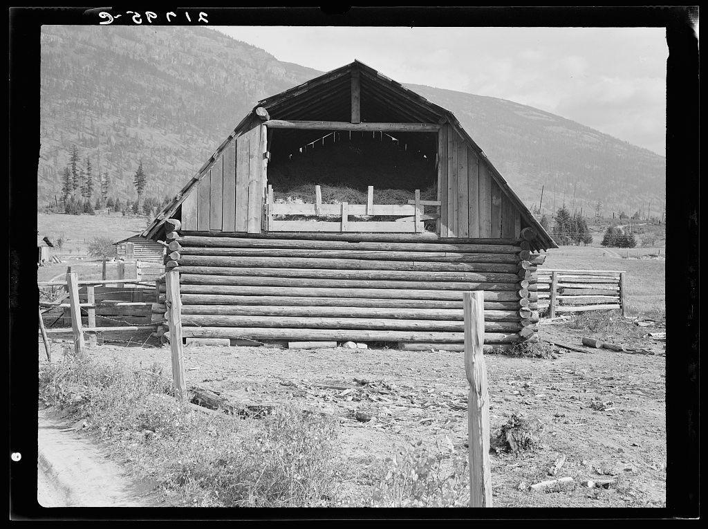The image is a black and white photograph depicting an old log barn situated in the countryside. The barn has a steep, pitched roof with open sides on two of its longer walls, allowing for ventilation or viewing purposes. It appears to be constructed from horizontal logs placed atop one another without any visible nails holding them together. A fenced enclosure made up of similar wooden logs surrounds the structure, suggesting that it may serve as an animal shelter or storage area.

The barn is situated in a field with sparse grass and dry soil, indicating arid conditions typical of certain rural regions. In the background, rolling hills covered with trees rise from the landscape, creating a backdrop for this pastoral scene. The overall setting suggests historical agricultural use, possibly within a region known as Boundary County, Idaho.

There are no people or animals visible in the photograph; it focuses on showcasing the barn and its immediate surroundings. This image is associated with FSA (Farm Security Administration) borrower plans to develop dairy ranches, indicating that this rural area may have been considered for agricultural expansion during the time when such photographs were taken by Dorothea Lange.

The specific details about general caption 56 can be found at: https://images.loener.nl/DorotheaLange/full/6495/6495b836ba25398d90c501b6.jpg.