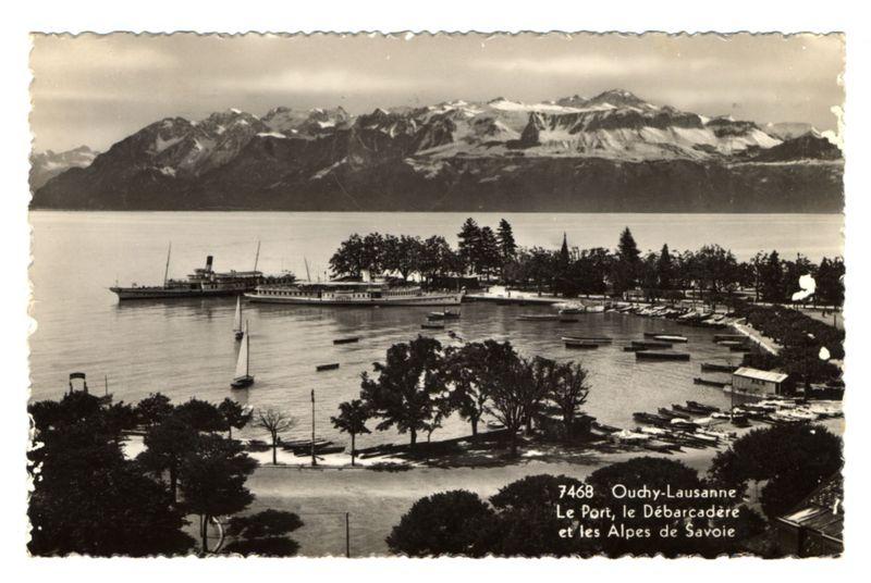 The image depicts a historical photograph of Lake Geneva, showcasing the port and harbor area in Oudry-Lausanne. The black-and-white photo captures several boats on calm waters with mountains rising majestically in the background. Prominent among them is a large steamship docked at the pier, surrounded by smaller sailing vessels. Onshore, there are trees lining the waterfront, some of which appear to be situated within what looks like parkland or gardens. A few buildings and structures can also be seen near the water's edge.

The photograph bears text in French on its right-hand side: "7468 Oudry-Lausanne Le Port, le Débarcadère et les Alpes de Savoie," which translates to "(Postcard number) 7468 Lausanne-Oudry Harbor and the Alps of Savoie." This suggests that this image is part of a postcard collection documenting various views from or associated with Lake Geneva in Switzerland, specifically highlighting its port area as well as connections to nearby Alpine regions. The text indicates an official attribution likely intended for postal use during the early 20th century when such images were popular souvenirs among travelers and collectors.

The overall ambiance of this image evokes a sense of historical charm and travel nostalgia, reflecting how scenic Swiss locales like Lake Geneva would have been perceived by visitors from other parts of Europe or even further back in time.