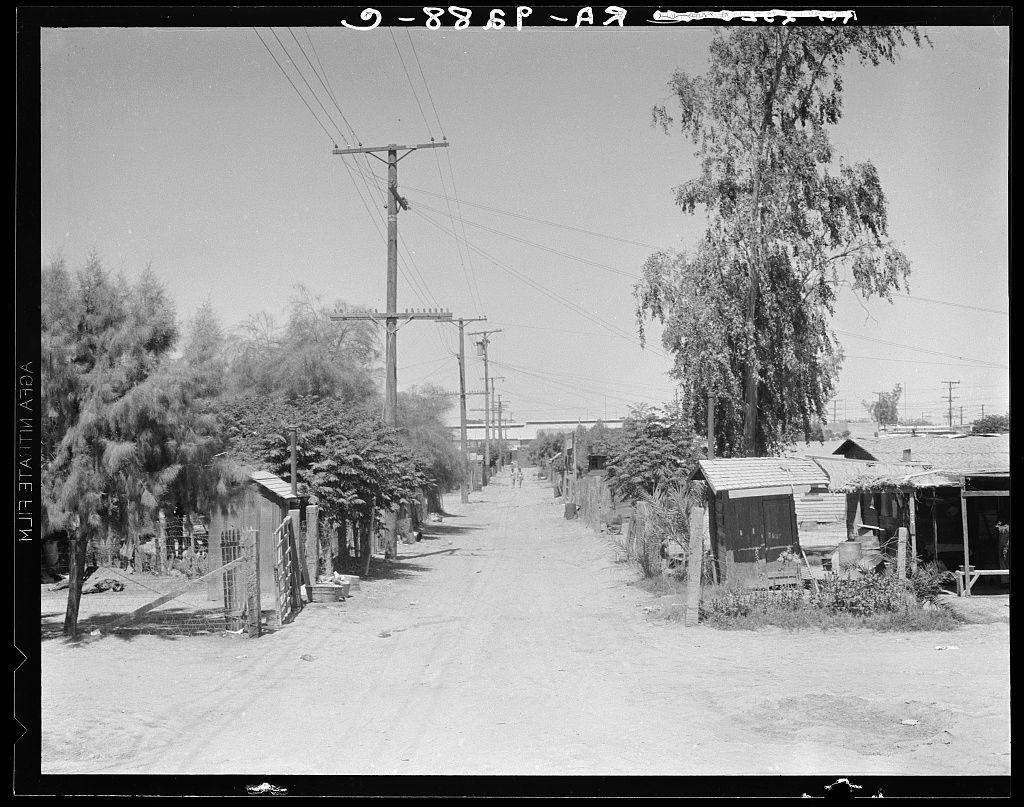 The image is a black and white photograph depicting an outdoor rural scene. It shows what appears to be a dusty, unpaved road flanked by simple wooden structures on both sides. These appear to be makeshift houses or sheds with sloped roofs, possibly indicating temporary housing conditions.

On the left side of the path, there are small trees or shrubs and some scattered debris near the buildings. The middle ground reveals more dilapidated constructions made from various materials such as wood and metal sheeting, suggesting that these structures might be used for living quarters but lack formal infrastructure like plumbing or electricity.

The right side shows similar conditions with additional wooden sheds and what seems to be a small fence barrier in front of one structure. There are electrical poles lining the road, indicating some level of utility service provision despite apparent neglect.

In the background, beyond the makeshift structures, there is an expanse of open sky and sparse vegetation including taller trees or bushes that extend into the distance. The overall impression is that of a semi-arid landscape with little sign of active industry or modern development in this area at the time when the photo was taken.