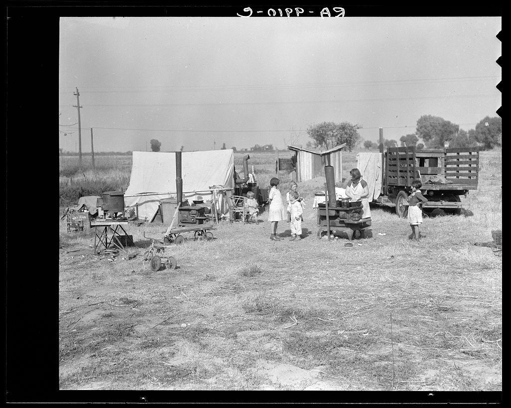 The image is a black and white photograph depicting a rural scene with several individuals, likely a family of migrants. There are six people visible: four adults and two children. They appear to be gathered around makeshift tents or temporary structures that provide some shelter from the elements.

In addition to the group of people, various household items and tools can be seen scattered around them, suggesting they may have been traveling with their belongings. These include a wooden cart loaded with what looks like logs or firewood, a metal water tank on wheels, and other equipment possibly used for agricultural work. 

The background shows an open field with sparse vegetation, indicating that the location could be in a rural farming area. A telephone pole stands tall against a clear sky, adding to the sense of being outside urban settings.

From the text overlaid at the top left corner "3-01P - 69", it seems like this photograph may have been taken by an organization or during some sort of survey from that year in Sacramento, California.