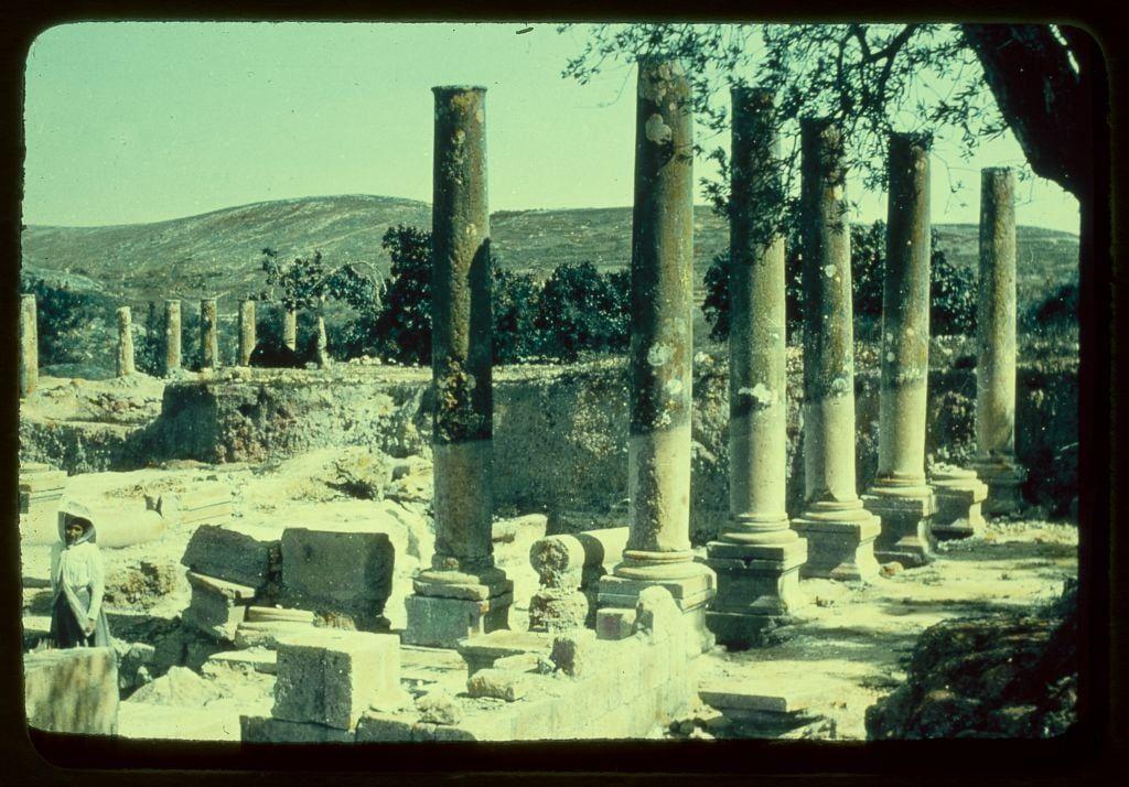 This image captures a scene of ancient ruins, likely taken in the mid-20th century judging by its appearance. The setting is an open area with scattered large stone blocks and columns that stand tall but show signs of weathering and age. Some of these column remnants are missing their capitals. In the background, rolling hills suggest a rural or semi-rural location.

A lone figure dressed in traditional attire stands to the left side of the photo, partially visible behind some fallen stones. The person appears contemplative, possibly observing the ruins with interest or reflection on history and time passed. 

The overall atmosphere is one of historical exploration, capturing remnants from an ancient civilization that has long since vanished.
