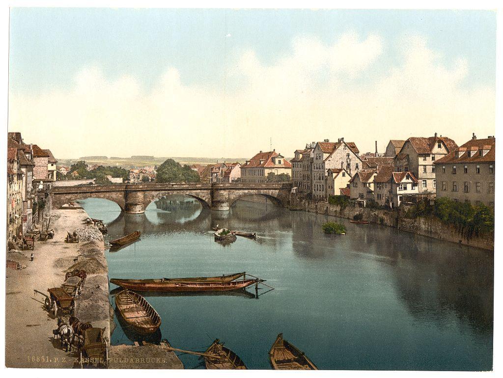 The image depicts a picturesque view of the Fulda River in Kassel, Hesse-Nassau, Germany. The scene captures an early 20th-century postcard with watercolor-like hues that give it an antique feel. A stone bridge stretches across the river, connecting two sides of the town adorned with traditional European architecture featuring red-tiled roofs and a mix of buildings in various states of repair.

Numerous boats are scattered throughout the calm blue waters; some appear to be canoes or small rowboats, while others seem larger, possibly serving as transport vessels. The left bank is bustling with activity: horse-drawn carts loaded with goods ready for delivery line up along the river's edge. A lone figure stands on a wooden platform by one of these carts.

The sky above is partly cloudy but brightens an overall pleasant atmosphere over the tranquil town scene, highlighting the bridge and its reflection in the calm waters below. The image appears to be part of a collection from around 1890 to ca. 1900, showcasing everyday life along one of Germany's rivers during that era.

Further context is provided by Fulda Bridge (Kassel), Hesse-Nassau, Germany between approximately 1890 and circa 1900 on a website called Views of Germany at the specified URL. This indicates an interest in historical photography from this region within Europe’s cultural landscape.