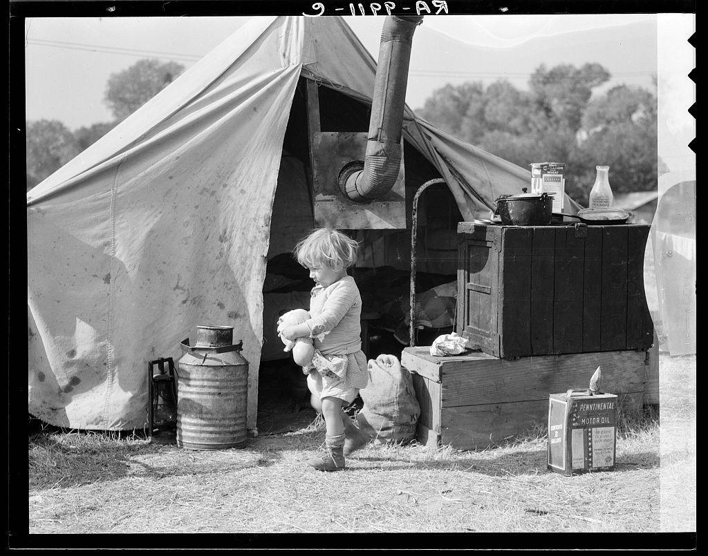 The image depicts a young child standing in front of a makeshift dwelling composed of various materials. The structure appears to be an improvised shelter, possibly used by migratory workers or during times of hardship. It has a large canvas-like covering supported by what looks like wooden beams and other miscellaneous items repurposed for construction.

Adjacent to the tent are several objects that suggest daily living activities: a metal canister with a lid, likely containing foodstuffs; a container labeled "Pennington's Motor Oil," suggesting transportation or mechanical work nearby; a milk churn, indicating attempts at domestic chores like cooking or cleaning; and a wooden barrel, possibly for storage of goods.

The child is wearing light-colored clothing suitable for cooler weather. They are holding what appears to be a doll in one hand while resting their other arm on the ground. The environment around them seems rural with trees visible in the background under an overcast sky, suggesting this campsite might not have access to modern amenities like electricity or plumbing.

The black and white nature of the photograph adds historical context, indicating that it may be a vintage image from when such living conditions were more common for certain populations. The overall scene conveys resilience amidst challenging circumstances.