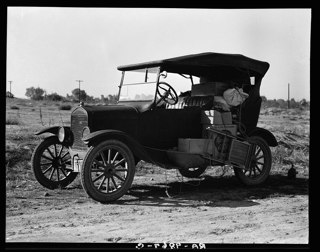 The image displays a vintage automobile parked outdoors on an unpaved surface. The vehicle appears to be from the early 1900s, characterized by its prominent front grille and headlamps mounted directly above each other. It features spoked wheels typical of that era's design. A canvas top is visible in place of a traditional roof, suggesting it may have been designed for leisurely drives or was modified to be more portable.

Inside the car, various items are loosely arranged on seats, implying either travel or temporary storage during transit. The surrounding environment includes sparse vegetation and utility poles stretching into the distance under an open sky with no visible structures close by. This setting suggests a rural area possibly used for agricultural purposes.