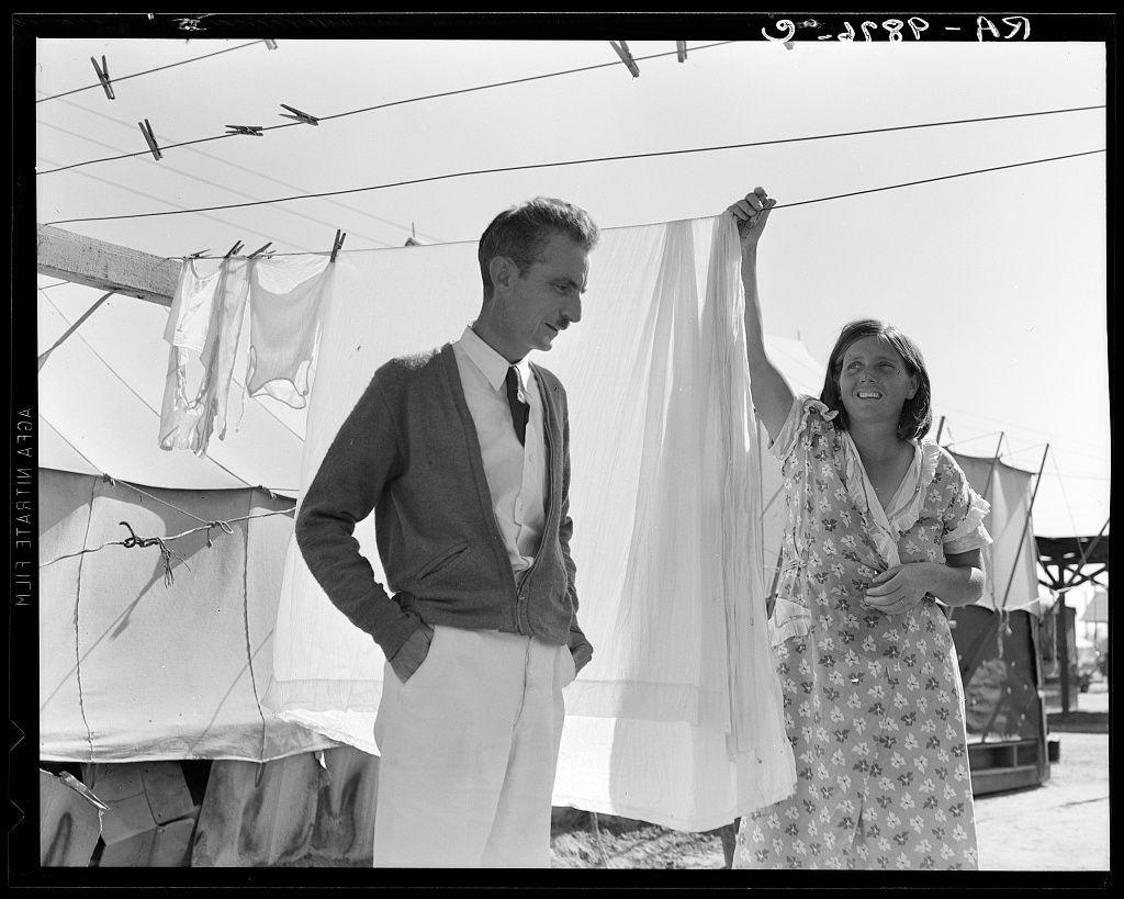 The image is a black and white photograph depicting two individuals, one male and one female, standing outdoors near an open-air laundry line. The man appears to be wearing business casual attire consisting of a cardigan sweater over a shirt with rolled-up sleeves, trousers, and shoes. He has his hands in the pockets and is smiling down towards the woman's actions.

The woman, possibly younger than the male individual, is dressed in what seems like a patterned summer dress or housedress with short sleeves and an open neckline. She holds up a piece of white fabric on a clothesline using her right hand while holding another similar garment in her left hand close to her chest area. Her expression suggests she is engaged in conversation with the man, laughing.

The surroundings indicate they are near some kind of temporary housing or tent structures commonly associated with migrant camps during the early 20th century. The laundry line has several clothespins attached and garments hanging on it, further supporting this setting as one that accommodates individuals possibly living temporarily due to migration work patterns typical in California's agricultural regions.

Additional information about this image can be found from Tom Collins, manager of Kern migrant camp, talking with one of the members.