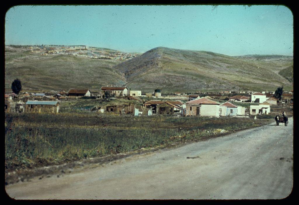 This image depicts a rural landscape featuring an assortment of small, modest buildings with varied roofing styles and colors. The architecture suggests either historical significance or simple construction methods common in pastoral settings. A dirt road runs parallel to the viewer's perspective on the right side, suggesting accessibility for travel but indicating that modern infrastructure is not prevalent here.

In the background stands a rolling hillside covered with sparse vegetation under clear skies, which gives an impression of arid climate conditions typical of semi-desert regions or Mediterranean climates in general. The absence of lush greenery or expansive trees reinforces this observation.

The image has a vintage quality, likely dating from mid-20th century judging by the color saturation and film grain visible on it. It is part of a collection named "Northward from Jerusalem," which suggests that these photos might be documenting geographical sites relevant to biblical history such as Nazareth or other related locales in Israel.

The photo was taken under Matson Photo Service, an organization known for its archive of photographs capturing landscapes and historical events between the 1930s through until approximately the mid-20th century. This image is particularly notable because it's associated with Esdraelon which could be a specific region within Israel  [...]