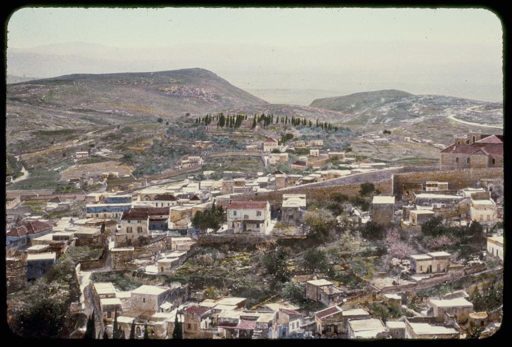 This image depicts a panoramic view of the Nazareth landscape as seen from an elevated vantage point. The area is densely populated with buildings, displaying various architectural styles and construction materials such as stone, brick, and tiled roofs in shades of red and orange. Amidst these structures are patches of greenery and cultivated fields, hinting at agricultural activity within or around the town.

In the background, rolling hills rise against a muted sky that fades into gentle mountain ranges further back. The landscape is characterized by an overall earthy palette with hints of verdant hues in selected areas like trees and vegetation on the hilltops. There's no visible modern infrastructure such as roads or vehicles, which lends to this being possibly from several decades ago.

The image suggests a historical context through its representation style - reminiscent of mid-20th-century photography that captures both the environment and community with an air of authenticity and simplicity before significant urban development took place in similar regions. This scene is part of 'Northward From Jerusalem,' as noted, which indicates geographical or biblical significance to those familiar with Christian narratives.

The specific caption reference suggests a connection to religious texts – likely referencing Jesus' "Spirit will drive them out" prophecy from the New T [...]