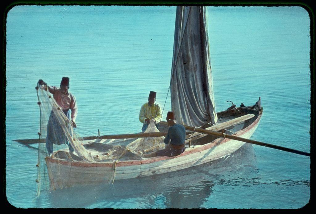 The image captures a traditional scene of fishermen on the Sea of Galilee. The focus is on three individuals aboard a small sailboat with one mast and an unfurled sail, which appears to be partially caught in some wind but not fully hoisted for sailing purposes. Two men are standing while handling fishing nets; they wear conical hats typically associated with Middle Eastern fishermen. One man sits at the stern of the boat wearing similar headgear. The sea is calm with a gentle reflection on its surface, indicative of clear weather conditions.

The colors in this image have muted tones, likely due to aging or camera processing techniques common during the late 20th century when it might be sourced from 'Around the Sea of Galilee.' This photo was taken by Matson Photo Service as part of their collection documented between approximately 1950 and 1977.