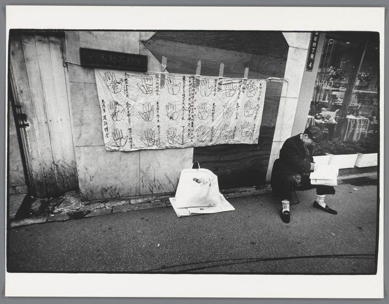 The image is a black and white photograph that captures an urban street scene in Japan. It features a person sitting on the pavement, engrossed in reading a newspaper spread out before them. The individual appears to be wearing dark clothing with a hat, as well as patterned socks or boots.

In front of this seated figure stands another large bag filled with items, possibly belongings or goods for sale, placed against what seems to be an outdoor market stall's wall which is partially covered by a signboard reading "永和不動". This could suggest the setting involves commerce and street vendors. The surrounding area shows other elements like windows reflecting city life, indicative of nearby buildings.

Prominently displayed on this wall are numerous hand prints or palm impressions accompanied by Chinese characters arranged in rows, possibly for artistic expression, advertisement, or communication purposes. These impressions may represent a form of social commentary or personal expressions from various individuals who have come to leave their mark behind at the site.

The overall atmosphere conveyed is one of everyday life and human interaction within an urban context. The use of black-and-white photography lends it a timeless quality, focusing on contrasts between light and shadow without color distraction.