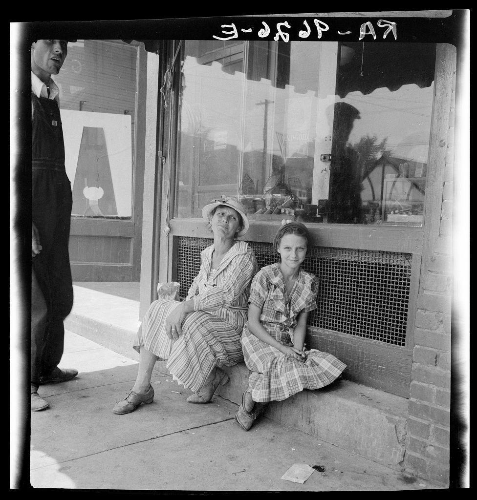 The image is a black and white photograph depicting two individuals sitting outside of what appears to be an establishment, possibly a store or diner. The person on the left seems male wearing overalls with shoes and holding something in his hand while gazing away from the camera. He stands beside another individual who sits next to him on a ledge.

The seated individual is dressed in a striped dress with buttons down the front, has their legs crossed at the ankles, and appears relaxed or contemplative. The person's right foot extends outward onto the sidewalk, which looks aged with some cracks visible. They have shoes that look worn but sturdy.

Next to this seated figure sits another young girl who is also in a striped dress, possibly plaid, showing a pattern of dark lines on lighter fabric. She has her legs spread out and appears focused or engaged in something at her feet. Her facial expression suggests she's either listening intently or perhaps absorbed by what’s happening around.

The background reveals the interior through large windows with metal mesh screens, suggesting that it could be during business hours as reflections indicate activity within. The exterior environment is bright enough to cast clear shadows on the ground and sidewalk, indicating daylight conditions.

Above them are handwritten inscriptions in white ink or paint, potentially identifying a date [...]