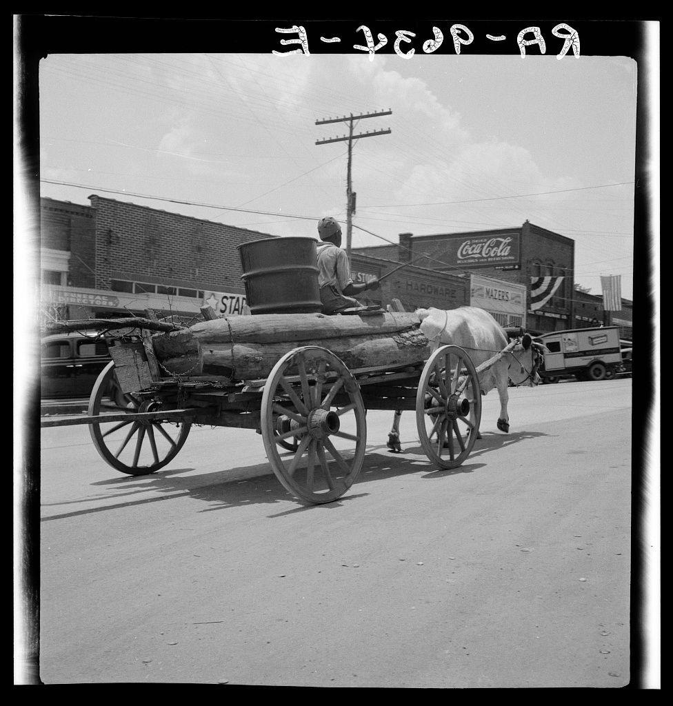 The photograph is a black and white image depicting an urban street scene from the early 20th century. It features a horse-drawn wagon, with a man sitting on top of it; he appears to be driving or steering the vehicle as they travel down what seems like a paved road.

In the background, we can see various commercial buildings including one that is prominently displaying an advertisement for Coca-Cola and another bearing signs such as "Star" and "Hardman Hardware." The presence of these business names suggests this might have been taken in Birmingham or Montgomery. An American flag is also visible on a pole behind the horse-drawn wagon, adding to the historical context.

The vehicle itself has large wooden wheels with metal spokes and appears functional rather than decorative. There are barrels attached at various points along its sides, which could be for carrying goods related to commerce during that era. The road looks somewhat busy given there's another car in the background traveling on a separate street or lane parallel to where the horse-drawn wagon is moving.

The photograph also contains handwritten text and numbers across the top right corner of it: "3-20P-'49," which likely indicates when this photo was taken (March 1949) as well as its internal numbering within a collection. The context provided suggests that Dorothea Lange, known for her work during the Great  [...]