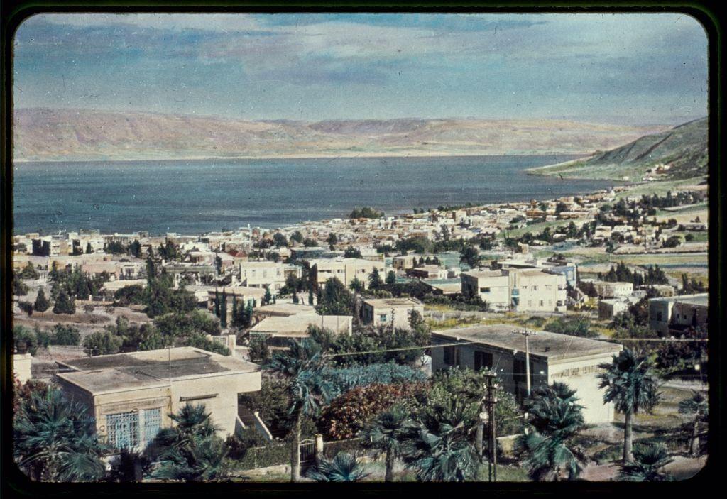 This image depicts a panoramic view of a town located beside the Sea of Galilee, with a backdrop of arid hills. The urban area is densely packed with buildings featuring flat and slightly sloping roofs, indicative of residential architecture common in Mediterranean climates. Numerous palm trees are scattered throughout the landscape, suggesting an attempt to cultivate greenery within this seemingly dry environment.

The town extends from the foreground where modern structures can be observed down towards a larger cluster of older-looking houses along the shore, with buildings varying in size and design but generally presenting a modest aesthetic. The sea itself is vast and calm, stretching out into the distance until it meets a range of brownish hills that fade gradually against a partly cloudy sky.

The photograph appears aged, showing signs of yellowing edges around the frame which could indicate its age or exposure to light over time. It seems like an archival image captured by Matson Photo Service during a period between 1950 and 1977 as per Around the Sea of Galilee: Tiberias from the west between 1950-1977, suggesting historical significance.

The picture is framed in what appears to be cardboard or similar material with rounded edges. The image has a vintage quality which may have implications on its authenticity and preservation status.