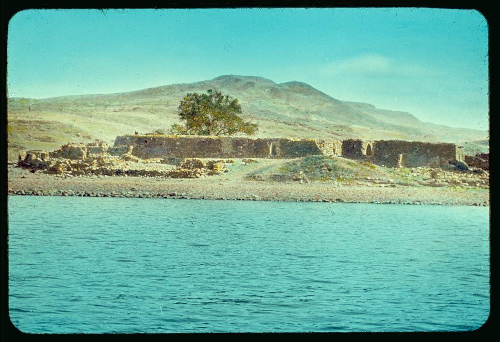 The image displays an ancient stone structure situated beside a serene body of water, likely the Sea of Galilee given its historical and geographical context. The remains appear weathered yet sturdy with visible walls that suggest once-formidable fortifications or perhaps religious significance in past centuries. A solitary tree stands within what could have been courtyard spaces among these ruins. In the backdrop, gentle hills rise against a clear sky indicative of arid climate conditions typical for this region. This tranquil landscape is framed by water on one side and land with rolling terrains extending into the distance behind it.