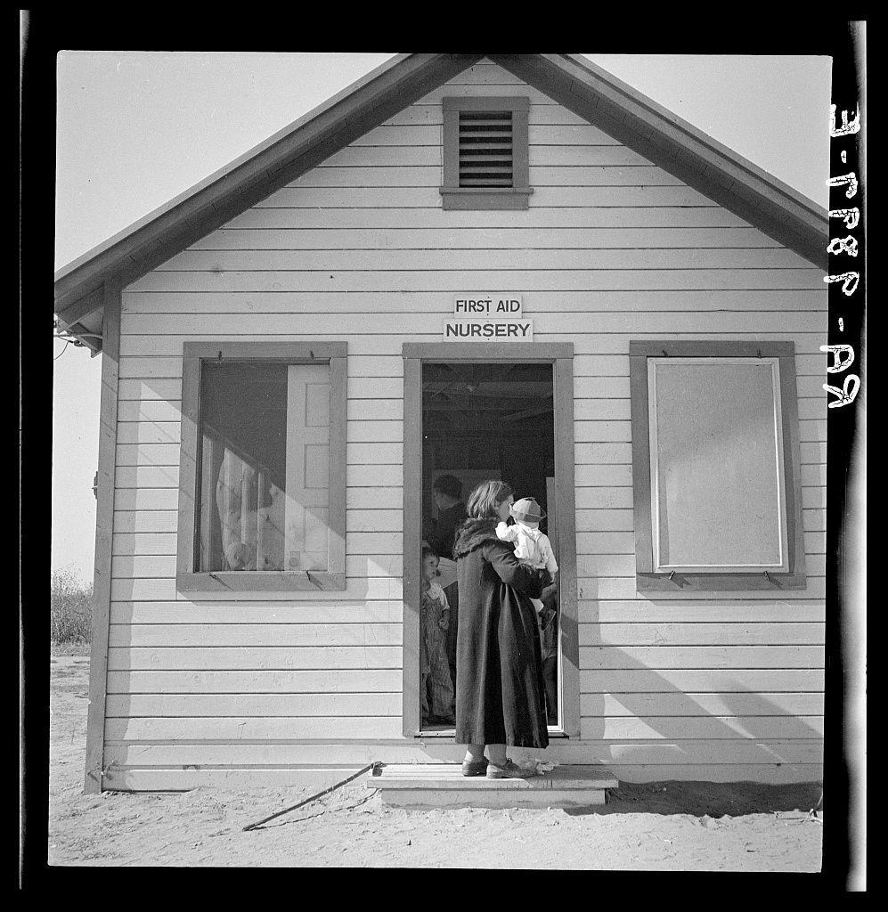 The photograph captures a modest wooden building with the inscription "FIRST AID NURSERY" above its entrance. The structure is simple, featuring white siding and has two visible windows; one with curtains partially drawn inside, suggesting privacy or protection from outside elements. To the right of the nursery's doorway stands an adult female figure holding what appears to be a young child in her arms, possibly awaiting entry into the building for medical attention or childcare services. The surrounding area is barren and sandy, indicating this might be part of a larger campsite or settlement with minimal vegetation around. Given its context as referenced from "Nursery Kern Migrant Camp," it's likely that this was taken during a time when migrant workers needed such facilities to care for their children while they worked in the fields nearby. The image is black and white, which adds an air of historical significance or nostalgia, typical of photographs dating back several decades ago.